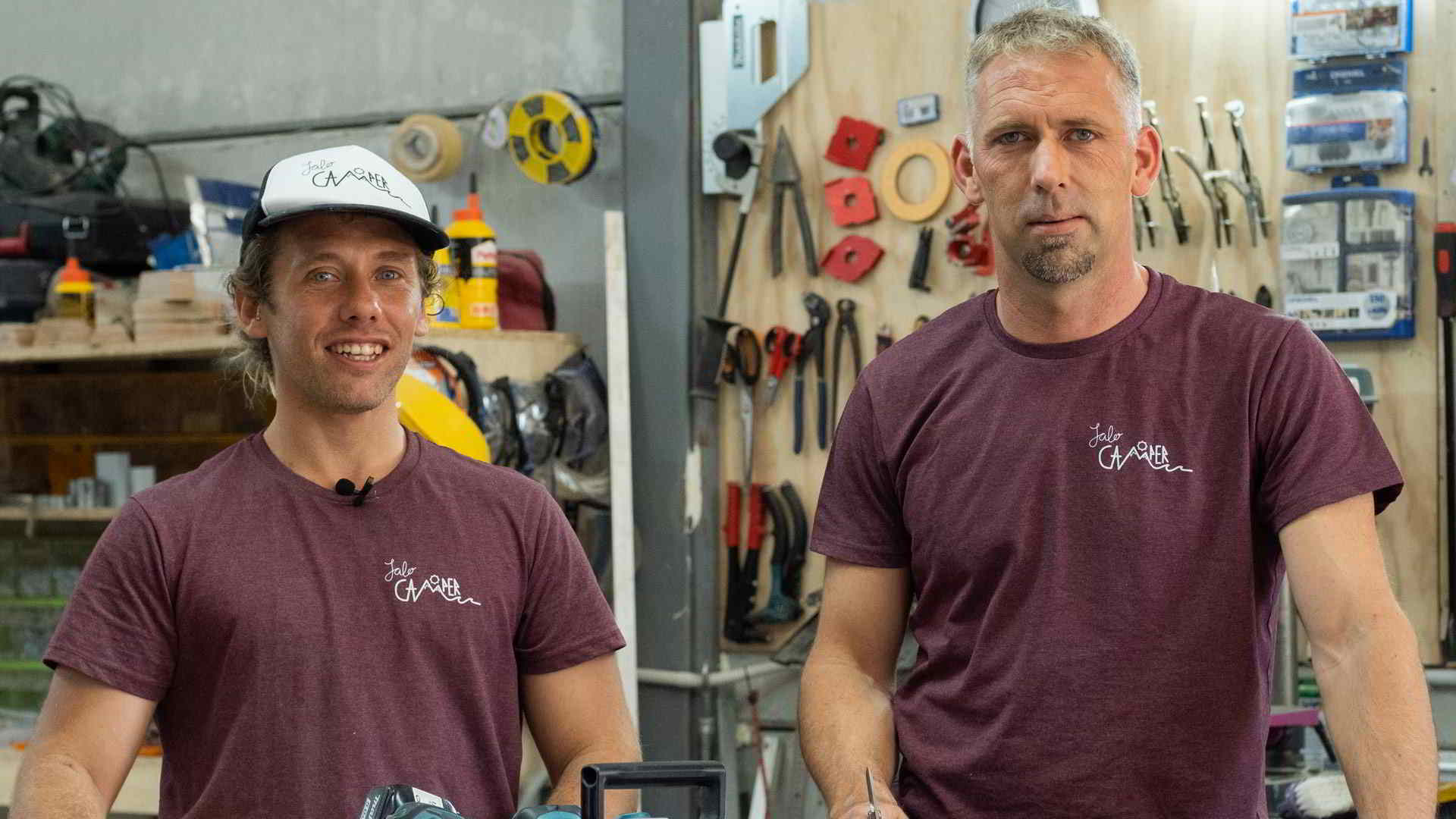 Dos miembros del equipo de Jalo Camper en su taller, vestidos con camisetas corporativas, rodeados de herramientas de carpintería y camperización.